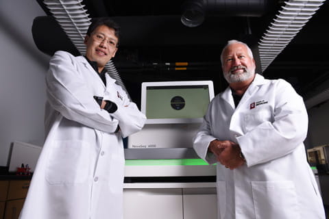 two scientists in lab coats stand with arms crossed in front of a lab instrument