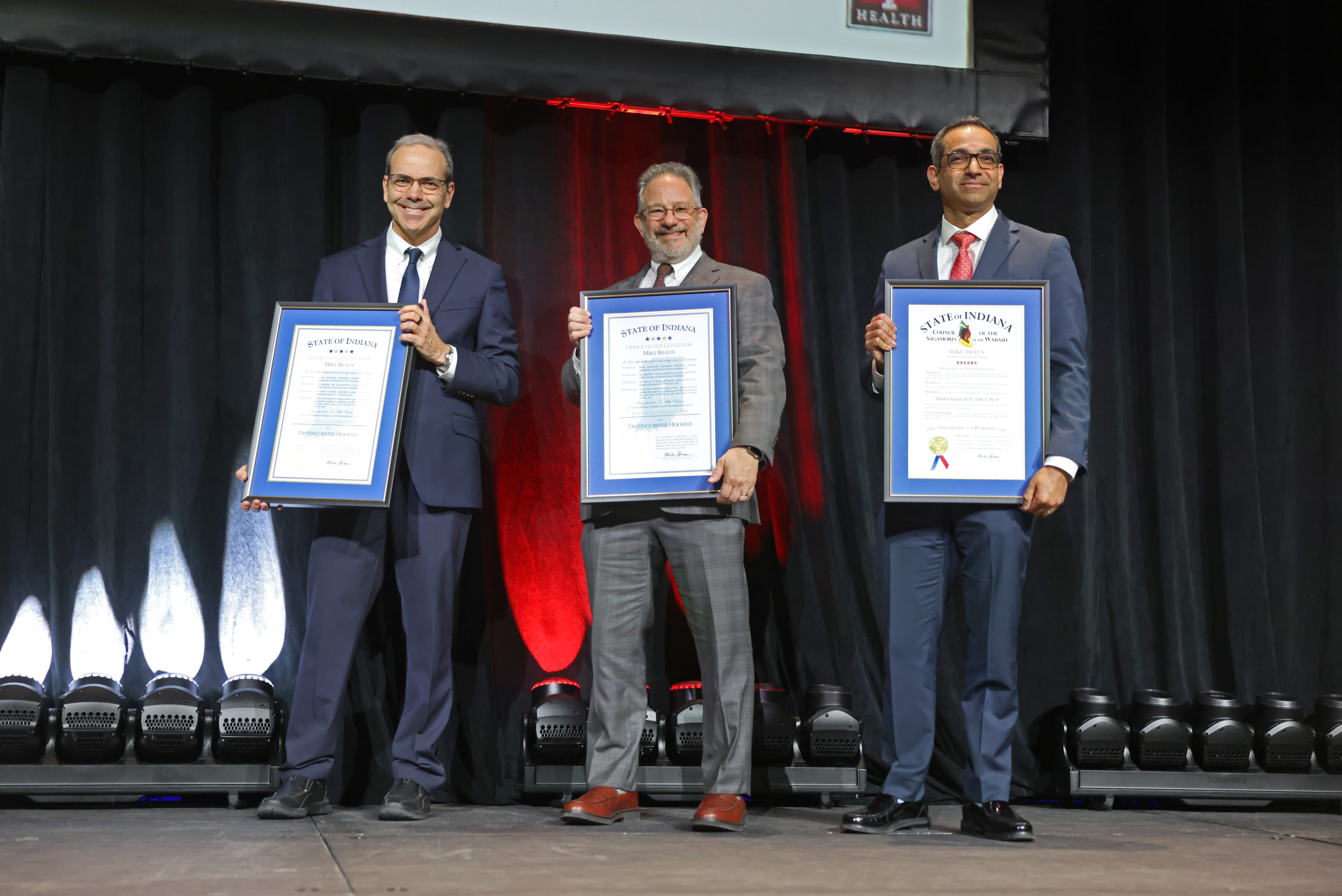 Three men in suits hold framed awards on a stage. 